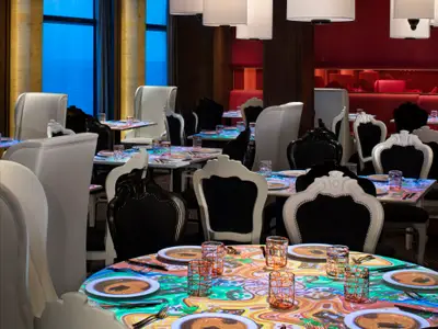 Ein elegantes Restaurant mit vielen leeren Tischen, die mit bunten, animierten Lichtprojektionen dekoriert sind. Schwarze und weiße Sessel stehen um die Tische. Im Hintergrund sind rote Sitzbänke und ein blauer Lichteinfall zu sehen.