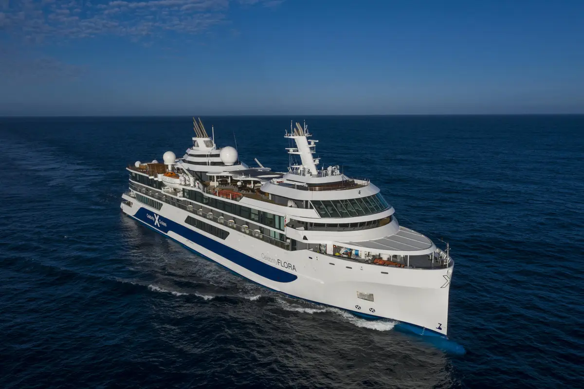 Luftaufnahme des Expeditionsschiffes Celebrity Flora auf blauem Meer unter klarem Himmel. Das Schiff ist weiss mit einem blauen Streifen am Rumpf und hat mehrere Decks sowie Antennen.
