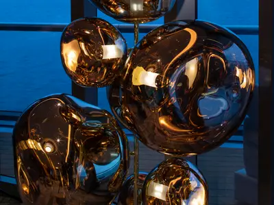 Nahaufnahme einer modernen Kunstinstallation, die aus mehreren glänzenden, organisch geformten, bronzefarbenen Kugeln besteht. Die Kugeln reflektieren interne Lichter und die blaue Umgebung des Wassers und des Himmels, die durch ein Fenster sichtbar sind.