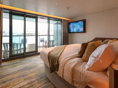 Moderne Kabine auf der Celebrity Flora. Großes Bett, viele Kissen, Balkontür mit Möbeln. Wand-TV, gestreifter Teppich. Warme Beleuchtung. Einladende Atmosphäre an Bord.