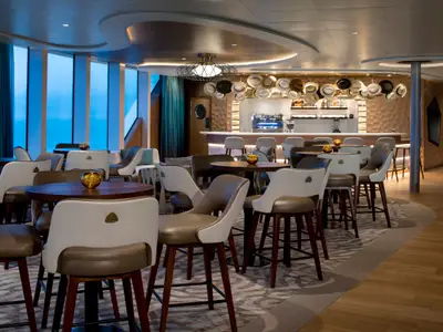 Elegante Bar auf der Celebrity Flora mit modernen Sesseln, Tischen und großen Fenstern mit Meerblick. Eine lange Theke ist mit einer einzigartigen Hutedekoration geschmückt.