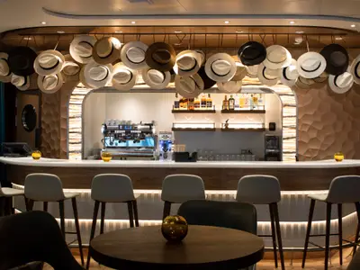 Moderner Bar-Bereich auf der Celebrity Flora. Eine weiße Theke mit Barhockern, einer Kaffeemaschine und Spirituosenregalen ist abgebildet. Über dem Tresen hängen dekorativ viele Hüte.