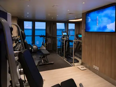 Moderner Fitnessraum an Bord der Celebrity Flora. Er verfügt über Panoramafenster mit Meerblick, zahlreiche Trainingsgeräte wie Laufbänder und Hanteln sowie einen großen Informationsbildschirm.