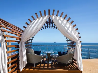 Zwei Sessel und ein Tisch unter einer Holzbogen-Pergola mit weißen Vorhängen auf dem Außendeck der Celebrity Flora. Blick auf das Meer mit einem fernen Schiff.