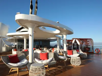 Sonniges Außendeck der Celebrity Flora mit weißer Überdachung, Lounge-Möbeln und roten Kissen. Holz-Cabanas und das Meer unter blauem Himmel sind zu sehen.