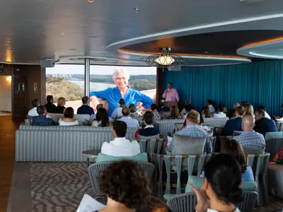 Gäste in einer modernen Lounge hören einer Präsentation auf einem großen Bildschirm zu.