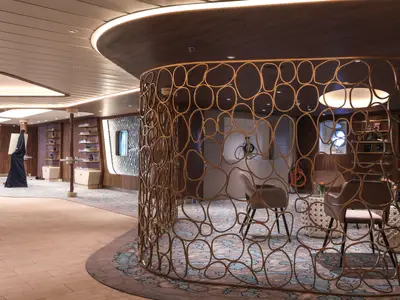 Interieur der Celebrity Flora: Moderner Lounge mit goldener, geschwungener Trennwand. Sessel, gemusterter Teppich, Holzwände und ein Bullauge sind sichtbar.