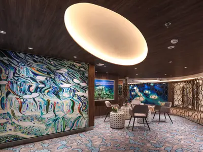 Lounge der Celebrity Flora. Mit großem, abstraktem Wandbild, Sesseln, Tischen, gemustertem Teppich und dekorativer Trennwand. Indirekte Beleuchtung.