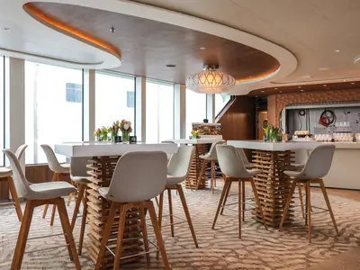 Heller Barbereich auf der Celebrity Flora mit hohen Holztischen, Barhockern und großen Fenstern. Blumen. Moderne Bar im Hintergrund. Geschwungene Decke mit Lampe.