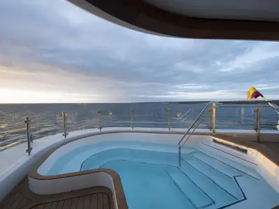 Ein Pool an Deck der Celebrity Flora mit Blick auf das Meer und eine Küstenlinie. Sonnenlicht erhellt den Horizont unter wolkigem Himmel. Eine Flagge weht am Heck.