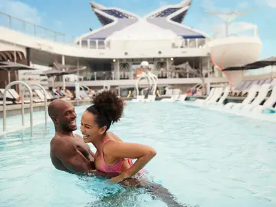 Ein lachendes Paar ist in einem Pool an Deck der Celebrity Flora. Im Hintergrund sieht man die Schiffsaufbauten, Sonnenliegen und andere Passagiere unter blauem Himmel.