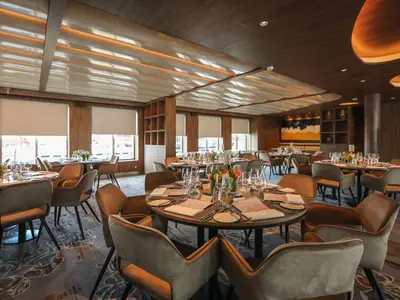 Restaurant der Celebrity Flora mit gedeckten Tischen, bequemen Stühlen und großen Fenstern. Helle Deckenbeleuchtung und Holzverkleidungen schaffen ein stilvolles Ambiente.