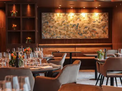 Das elegante Restaurant der Celebrity Flora. Gedeckte Tische mit vielen Weingläsern und Blumen, umgeben von Holzeinrichtung und einem Kunstwerk.