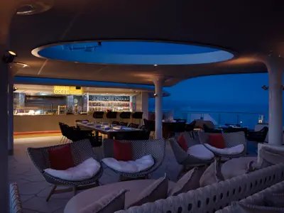 Abendansicht des Außendecks der Celebrity Flora: Bar, gedeckte Tische und Lounge-Sessel unter beleuchtetem Dach. Im Hintergrund das dunkelblaue Meer.