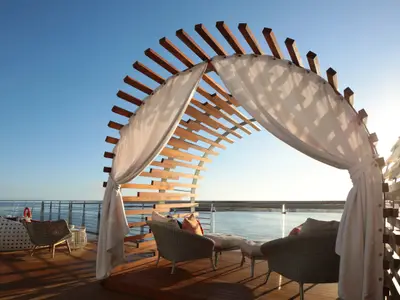 Loungebereich auf dem Außendeck der Celebrity Flora mit geschwungener Holzpergola, weißen Vorhängen und modernen Outdoor-Möbeln. Meerblick unter blauem Himmel.