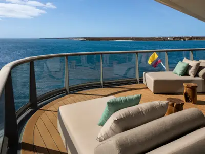Ein geräumiger Balkon mit Holzdeck und modernen Loungemöbeln. Im Hintergrund ist das blaue Meer und eine Küstenlinie unter einem klaren Himmel zu sehen. Eine kleine Kolumbienflagge ist am Geländer befestigt.