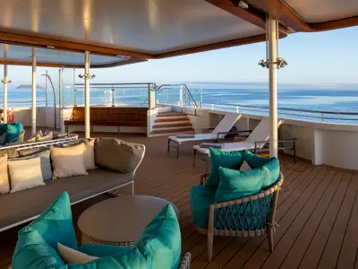Außendeck der Celebrity Flora mit Lounge-Möbeln und Sonnenliegen. Holzdeck mit Meerblick auf blaues Wasser und Insel am Horizont, sonnenbeschienen.