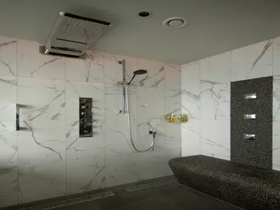 Eine moderne Duschkabine mit weißen Marmorfliesen und einem dunklen Mosaik-Akzentstreifen. Verschiedene Duschköpfe und Armaturen sind sichtbar. Ein gepolsterter Sitz befindet sich im Vordergrund.