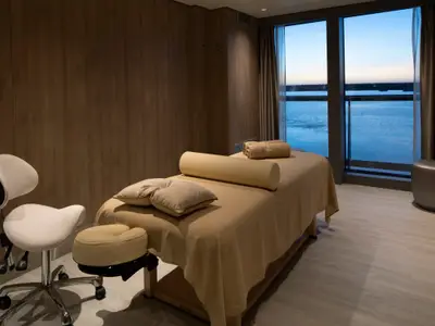Ein Spa-Raum der Celebrity Flora mit Massagebank und einem weißen Behandlungsstuhl. Große Fenster bieten einen weiten Blick auf das ruhige Meer bei Dämmerung.