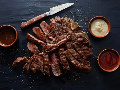 Ein appetitlich angerichtetes, in Scheiben geschnittenes T-Bone Steak auf einem dunklen Untergrund, begleitet von drei Schalen mit Saucen und einem Messer.