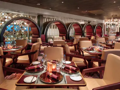 Elegantes Restaurant auf der Celebrity Flora mit gedeckten Tischen, beigen Stühlen und roten, halbrunden Sitznischen im Hintergrund. Stilvolle Deckenbeleuchtung ist erkennbar.