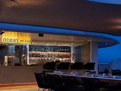 Eine gut beleuchtete Outdoor-Bar bei Abendstimmung. Ein Schild 'Ocean Grill & Bar' leuchtet hell. Die Bar ist mit zahlreichen Flaschen und Gläsern bestückt. Eine lange Tischreihe mit schwarzen Stühlen steht davor.