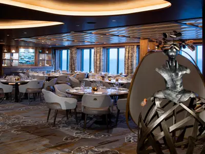 Geräumiges Restaurant der Celebrity Flora mit runden Tischen, bequemen Stühlen und Meerblick durch große Fenster. Eine auffällige moderne Skulptur steht rechts im Vordergrund.