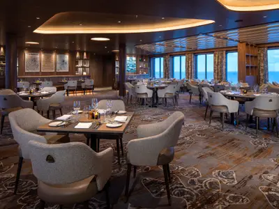 Elegantes Restaurant der Celebrity Flora. Viele gedeckte Tische, bequeme Sessel, Meerblick durch große Fenster. Holz, Teppich und dezente Beleuchtung.
