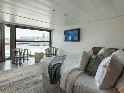 Helle Kabine auf der Celebrity Flora mit Doppelbett, Wand-TV und großem Fenster. Blick auf Hafen mit Kränen und Schiffen. Zwei Sessel und ein Beistelltisch stehen davor.