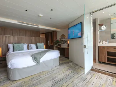 Die Innenansicht einer modernen Kreuzfahrtkabine auf der Celebrity Flora zeigt ein großes Bett, einen Flachbildfernseher und den Zugang zu einem offenen Badezimmer mit Holzmöbeln.