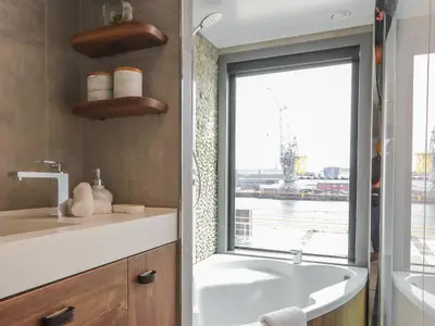 Modernes Badezimmer auf der Celebrity Flora mit Waschtisch, Dusche und Badewanne. Ein großes Fenster bietet Ausblick auf einen Industriehafen mit Kränen und Wasser.