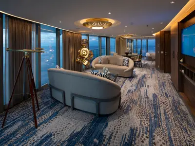 Innenansicht einer Suite der Celebrity Flora: Wohnbereich mit runden Sofas, Teleskop und Meerblick durch Fenster. Auffälliger Teppich in Blau-Brauntönen.