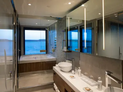 Modernes Badezimmer auf der Celebrity Flora: Badewanne am Fenster mit Meerblick, Glasdusche und Waschtisch mit Waschbecken und beleuchtetem Spiegel.