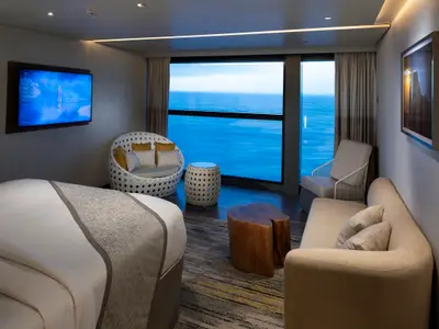 Helle Kabine der Celebrity Flora mit Panoramablick auf das Meer. Moderne Einrichtung: Bett, Sitzecke mit Sofa und Sessel, ein großer Fernseher an der Wand.