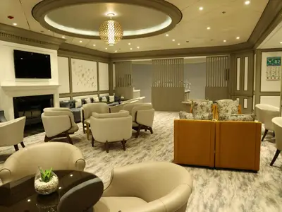Blick in die stilvolle Lounge der Celebrity Infinity. Moderne Sitzgruppen in hellen Farben, Kamin mit TV und goldene Pendelleuchte. Im Hintergrund weitere Bereiche und ein Notfallplan.