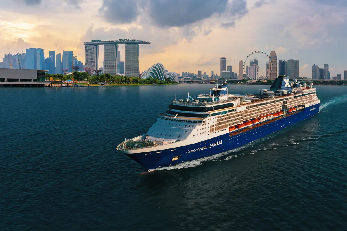 Ein blau-weißes Kreuzfahrtschiff mit dem Namen "Celebrity Millennium" fährt auf dem Wasser. Im Hintergrund ist die Skyline von Singapur mit bekannten Gebäuden wie dem Marina Bay Sands zu sehen.