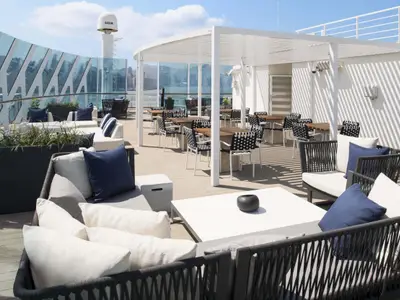 Sonniges Oberdeck der Celebrity Millennium mit modernem Outdoor-Lounge-Bereich und Essbereich. Helle Möbel, blaue Kissen, weiße Pergola. Im Hintergrund ist eine Stadtkulisse sichtbar.