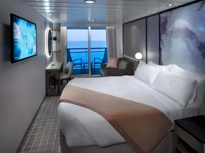 Moderne Kabine an Bord der Celebrity Millennium mit Doppelbett und Balkon. Durch die Glastüren ist der offene Ozean sichtbar. Ein Schreibtisch mit Stuhl und ein Sofa sind ebenfalls Teil der Einrichtung.