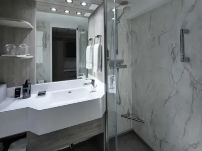 Ein modernes Badezimmer an Bord der Celebrity Millennium, ausgestattet mit weißem Waschtisch, Spiegel, und einer Duschkabine mit Marmoroptik und Chromarmaturen.