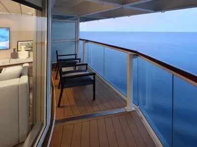 Blick aus einer modernen Kabine der Celebrity Millennium. Ein Balkon mit zwei Stühlen und Glasgeländer bietet weiten Meerblick. Innen sind ein Sofa und ein TV sichtbar.