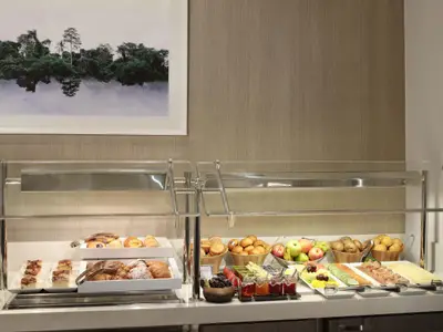 Ein Frühstücksbuffet mit einer Auswahl an Backwaren, frischem Obst, Müsli, Aufschnitt und Käse. Darüber hängt ein Bild einer tropischen Landschaft mit Spiegelung im Wasser.