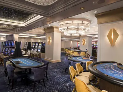 Szene im Casino der Celebrity Millennium: Blaue Spieltische mit Stühlen, dazu Reihen von Spielautomaten. Eine große Deckenleuchte und goldene Wandleuchten erhellen den Raum.