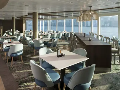 Helles Restaurant an Bord der Celebrity Millennium mit zahlreichen Tischen, blauen Stühlen und einer Bar. Große Fenster bieten Panoramablick auf das Meer und eine Skyline.