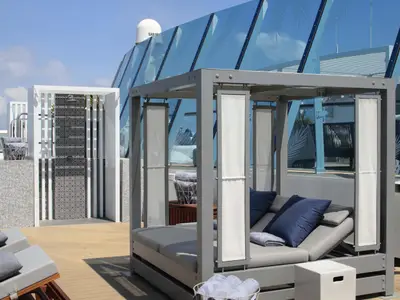 Sonniges Deck der Celebrity Millennium mit Außendusche und Tagesliege. Glasstrukturen und blauer Himmel im Hintergrund.