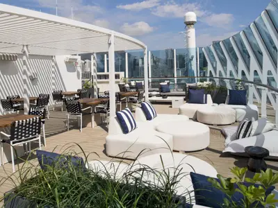 Ein stilvoller Außenbereich auf der Celebrity Millennium mit hellen Lounge-Möbeln, Esstischen unter einer Pergola und einem weiten Blick auf eine Stadtkulisse.