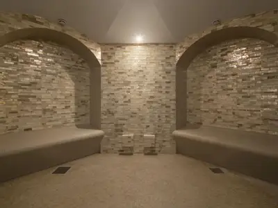 Blick in einen leeren Spa-Bereich oder Hamam. Zwei geschwungene, geflieste Sitzbänke stehen sich gegenüber, die von Bögen eingerahmt werden. Die Wände sind mit kleinen, rechteckigen Fliesen in verschiedenen Beige- und Brauntönen versehen, die im Licht schimmern. Ein helles Licht strahlt von der Decke, das sich in den glänzenden Fliesen widerspiegelt.