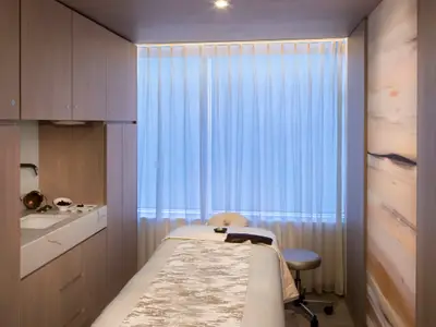Ein luxuriöser Spa-Behandlungsraum mit einer Massageliege in der Mitte, einem Waschbecken und Schränken auf der linken Seite und einem Fenster mit Vorhängen im Hintergrund. Eine dekorative Wand auf der rechten Seite.