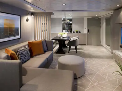 Moderner Wohnbereich einer geräumigen Suite mit L-Sofa, Esstisch, Minibar und TV. Graue Töne, orange Kissen, Wandbild. Heller Boden mit geometrischem Muster.