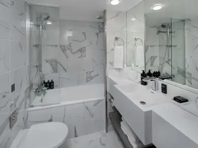 Ein modernes Badezimmer mit weissen Marmorfliesen an den Waenden und einer Badewanne mit Duscharmatur und Glaswand. Rechts befindet sich ein weisser Waschtisch mit schwarzer Seife und einem Spiegel. Das Licht ist hell und klar. 