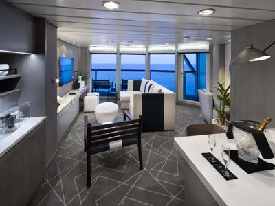 Eine luxuriöse Suite auf der Celebrity Millennium mit Wohnbereich, Meerblickfenstern und einer Bar. Das Interieur ist modern mit hellem Sofa, TV und geometrischem Teppichmuster.